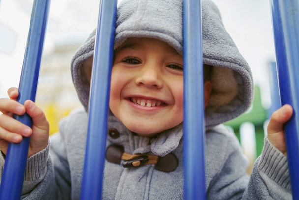 Portrait de petit garçon caucasien vêtu d'un manteau avec capuche sur la tête à travers les barres d'acier bleues souriant le jour d'hiver - Photo, image
