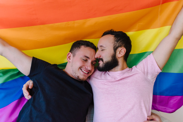 2人の白人同性愛者の男性が抱き合ってLGBTQの旗を掲げている写真。目を閉じてる. - 写真・画像