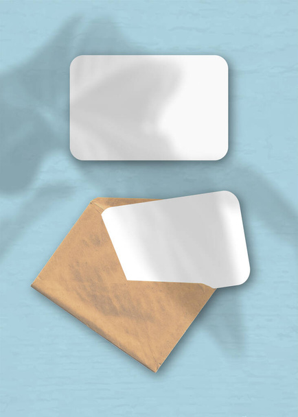 Ein Umschlag mit zwei Blättern strukturierten weißen Papiers auf blauem Hintergrund. Mockup-Overlay mit den Schatten der Pflanze. Natürliches Licht wirft Schatten von einer exotischen Pflanze. Vertikale Orientierung. - Foto, Bild