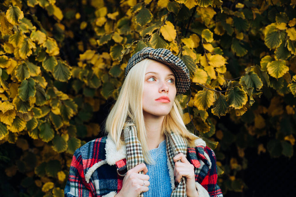 Beautiful romantic blonde girl standing at fall yellow autumnal leaves and looks dreamy. Beautiful femme wearing plaid scarf and cap in parisian style. Autumnal fashion concept. - Foto, afbeelding