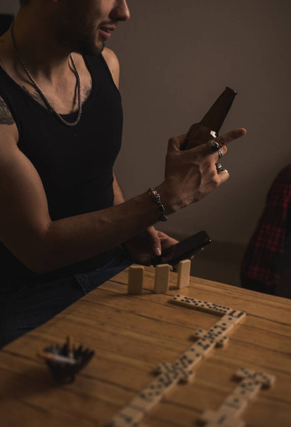 Rude young man playing dominoes with his friends, drinking beer while talking and laughing with his friends, on the table are dominoes, cigarettes, and in his hand he is carrying multiple rings - Fotó, kép