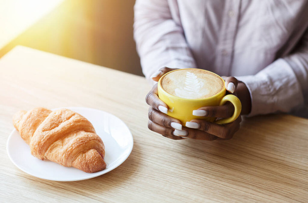 Vreedzame ochtend. Onherkenbaar Afro-Amerikaans meisje dat koffie drinkt met croissant aan tafel in gezellig café, close-up - Foto, afbeelding