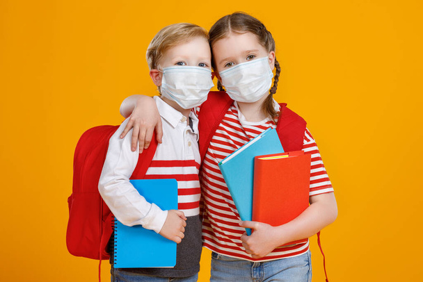 Niño y niña con máscaras que llevan libros de texto y se abrazan mientras van a la escuela durante la epidemia contra el fondo amarillo
 - Foto, imagen