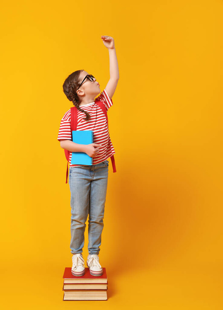 Πλήρες σώμα χαρούμενο κορίτσι με γυαλιά στέκεται σε στοίβα των εγχειριδίων και την αύξηση του βραχίονα, ενώ προσπαθεί να είναι υψηλότερη κατά τη διάρκεια των σπουδών σχολείο κατά κίτρινο backgroun - Φωτογραφία, εικόνα