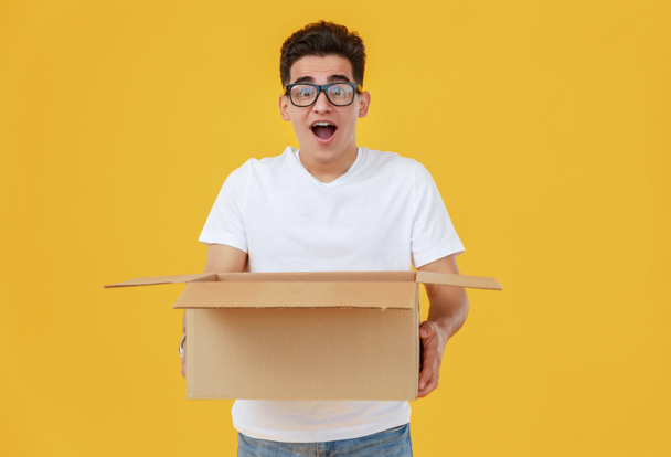 Hombre joven étnico sorprendido en gafas con caja de dibujos animados con boca abierta contra fondo amarillo
 - Foto, Imagen