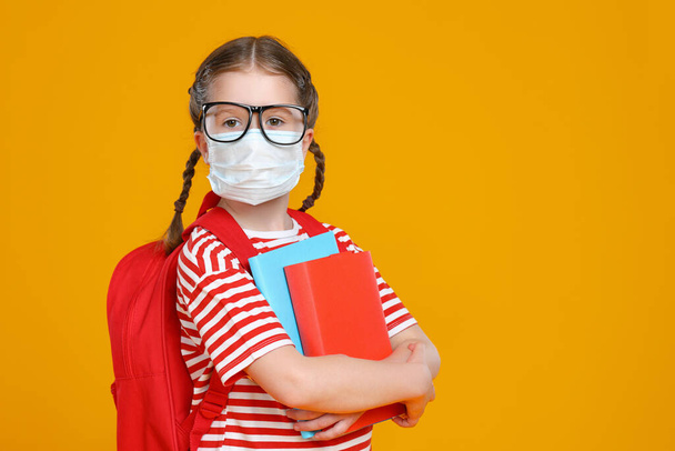 Έξυπνο κορίτσι με μάσκα και γυαλιά που αγκαλιάζει σχολικά βιβλία και κοιτάζει την κάμερα ενώ σπουδάζει στο σχολείο κατά τη διάρκεια πανδημίας ενάντια στο κίτρινο backgroun - Φωτογραφία, εικόνα