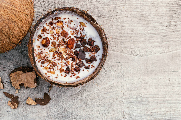 Coconut milk and granola made from oatmeal, nuts,dried fruits and grated chocolate in a coconut shell on a wooden background.oncept of healthy food and healthy desserts.Copy space for text - Fotoğraf, Görsel