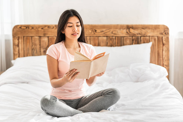 Азіатська дівчина читає книгу вдома сидячи на ліжку
 - Фото, зображення