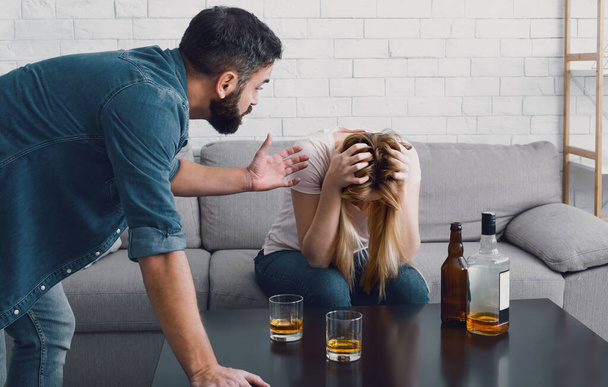 Drunken scandal. Woman holds her head and sitting on sofa, angry man swears with wife - Фото, зображення