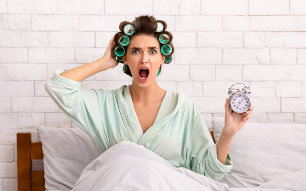 Mulher chocada segurando relógio sendo tarde sentado na cama dentro de casa
 - Foto, Imagem