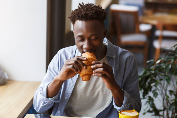 Modern életmód. Afrikai amerikai férfi croissant-t eszik ebédre a kávézóban - Fotó, kép