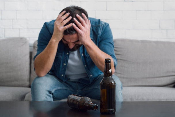 Smutný muž drží hlavu a sedí na gauči, prázdné láhve na stole, zblízka - Fotografie, Obrázek
