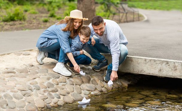 公園で小さな川で紙のボートで遊んでいる子供と彼の両親 - 写真・画像