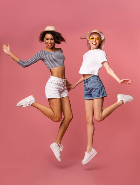 Summer Fashion. Two young girls in stylish clothes jumping on pink background - Foto, imagen