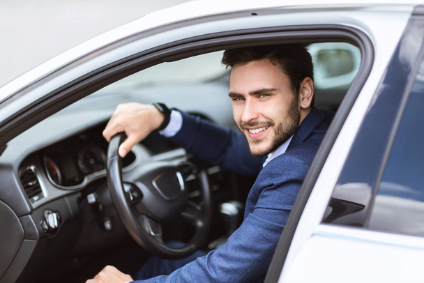 Handsome smiling entrepreneur sitting on drivers seat of his auto in city - Foto, imagen