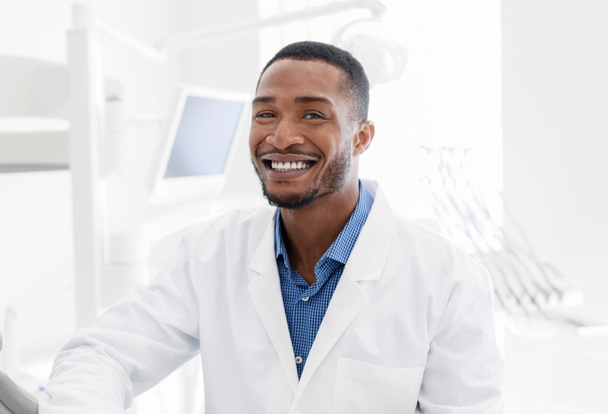 Knappe zwarte tandarts glimlachen over moderne tandheelkundige kliniek achtergrond - Foto, afbeelding