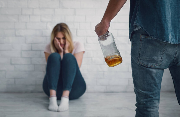 Hombre amenaza a mujer con botella en la mano, llorando mujer está sentado en el suelo
 - Foto, imagen