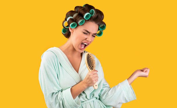 Νοικοκυρά με βούρτσα μαλλιών τραγουδώντας έχοντας διασκέδαση σε κίτρινο φόντο - Φωτογραφία, εικόνα