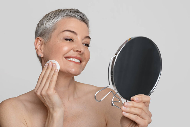 Schöne Frau mittleren Alters schaut in den Spiegel und putzt ihr Gesicht mit Wattebausch - Foto, Bild