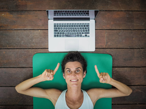 emotional woman lying on yoga mat on floor with laptop behind her head - Фото, зображення