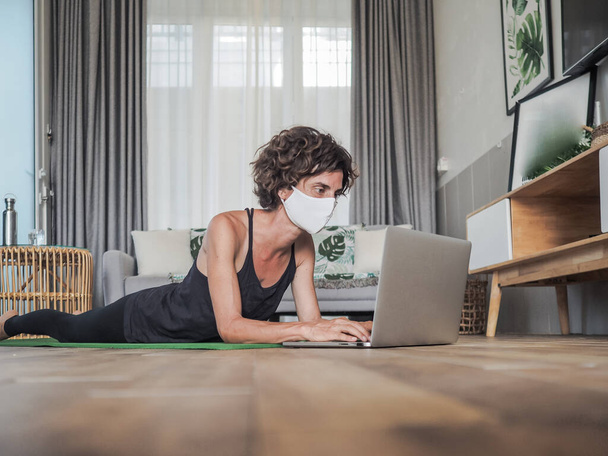 Maskeli kadın evde dizüstü bilgisayar kullanıyor yoga minderinde yatıyor. - Fotoğraf, Görsel