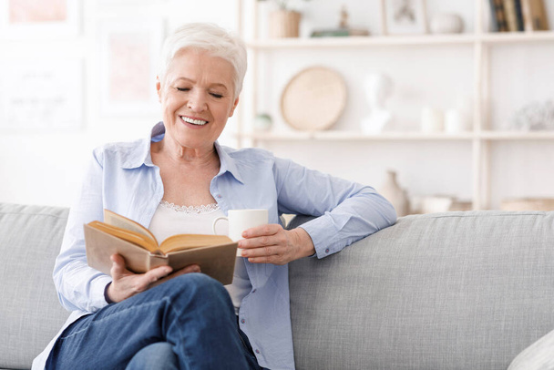 Zeit für den Ruhestand. Ältere Dame sitzt mit Buch auf Couch und trinkt Kaffee - Foto, Bild