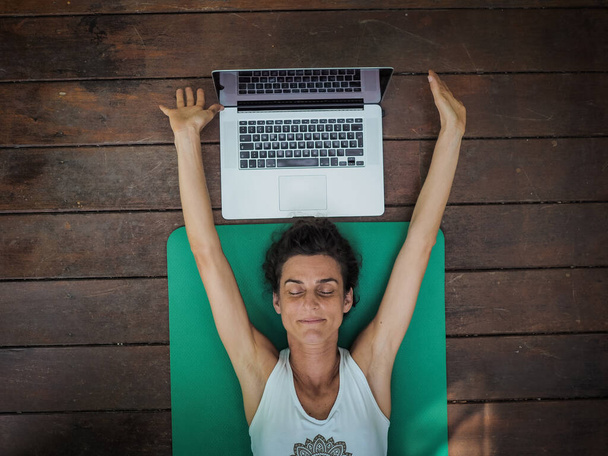 emotional woman lying on yoga mat on floor with laptop behind her head - Fotoğraf, Görsel