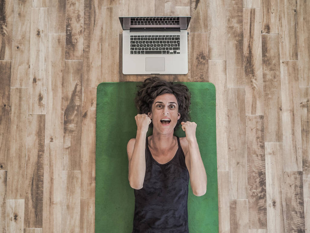 emotional woman lying on yoga mat on floor with laptop behind her head - Photo, Image