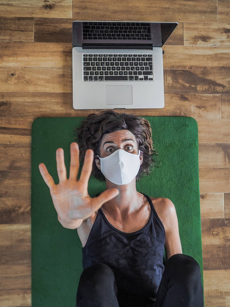 emotional woman in mask lying on yoga mat on floor with laptop behind her head - Фото, зображення
