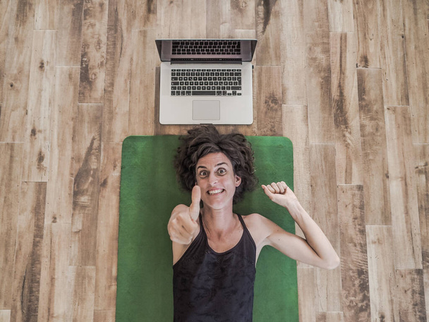 emotional woman lying on yoga mat on floor with laptop behind her head - Zdjęcie, obraz