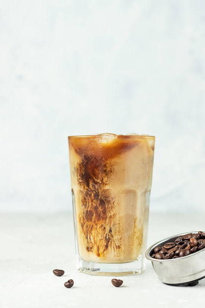 Iced coffee with milk in a tall glass with roasted coffee beans on light grey background. Summer refreshment drink. Selective focus. - Foto, afbeelding