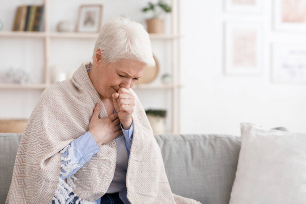 Άρρωστη ηλικιωμένη γυναίκα καλυμμένη με κουβέρτα που βήχει δυνατά στο σπίτι - Φωτογραφία, εικόνα