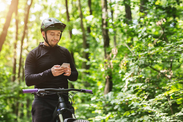 Молодий гонщик, стоячи в лісі, тримає мобільний телефон
 - Фото, зображення