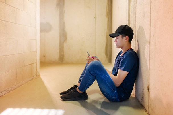 Joven sentado en el suelo en un pasillo o pasillo usando un teléfono móvil a la vista lateral
 - Foto, imagen