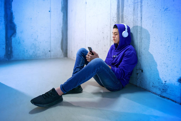 Menino adolescente sentado ouvindo música no chão em uma passagem de metrô ou comercial em um capuz azul quente e jeans em luz azul tonificada
 - Foto, Imagem