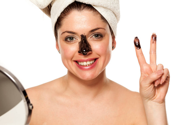 Joven hermosa mujer con toalla en la cabeza aplicando máscara negra en la nariz y mostrando los dedos
 - Foto, Imagen