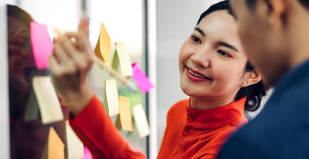 カジュアルな創造的な幸せな2つの笑顔アジアのビジネスの人々戦略分析とブレインストーミングを計画する人々は、現代のオフィスでガラス基板上のスタートアッププロジェクトのアイデアを共有するためにそれステッカーノートを置く - 写真・画像