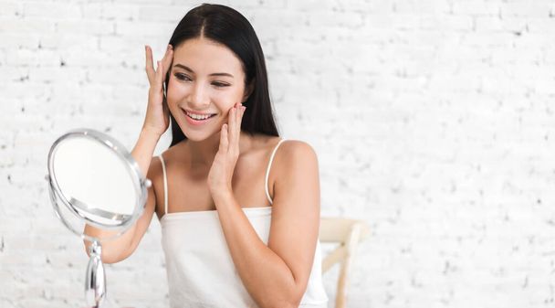 Smiling of young beautiful woman clean fresh healthy white skin looking at mirror.girl touching on her face with hand and applying cream at home.spa and beauty concept - Фото, изображение