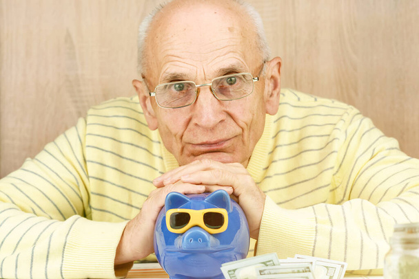 old man looks straight covering blue piggy bank slot - Foto, Bild