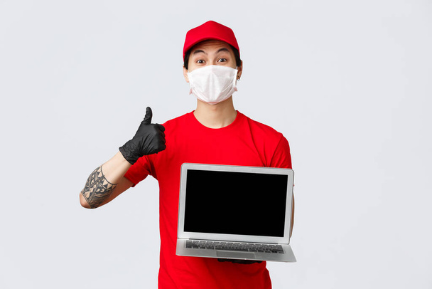 Good online service for food delivery or shopping. Cheerful courier in red cap and t-shirt, delivery guy holding laptop, wear medical mask, gloves, thumb-up, recommend contactless deliver of parcel - Foto, Imagem