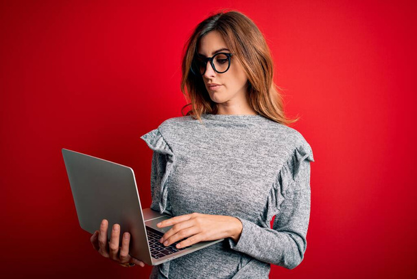 Młoda piękna brunetka businesswoman noszenia okularów za pomocą laptopa na czerwonym tle z pewnością wyraz na inteligentnej twarzy myśląc poważnie - Zdjęcie, obraz