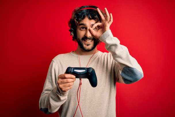 Joven jugador hombre con el pelo rizado y la barba jugando videojuego usando joystick y auriculares con la cara feliz sonriendo haciendo signo bien con la mano en el ojo mirando a través de los dedos
 - Foto, imagen