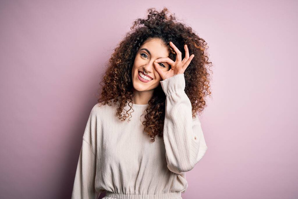 Beautiful woman with curly hair and piercing wearing casual sweater over pink background doing ok gesture with hand smiling, eye looking through fingers with happy face. - Foto, immagini