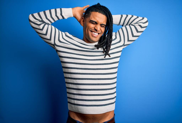 Young handsome african american afro man with dreadlocks wearing casual striped sweater relaxing and stretching, arms and hands behind head and neck smiling happy - Фото, изображение
