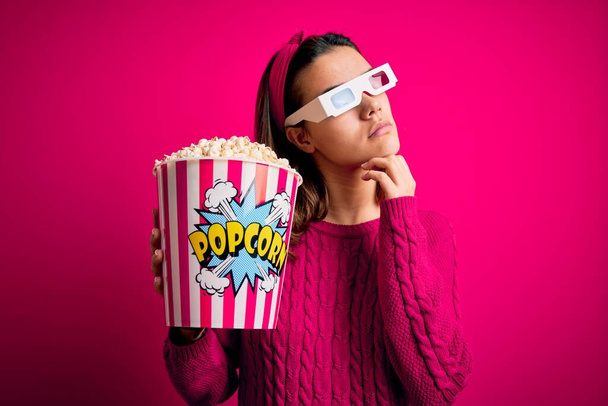 Junge schöne Mädchen, die Film mit 3D-Brille essen Box mit Popcorn mit der Hand am Kinn denken über Frage, nachdenklichen Ausdruck. Lächeln mit nachdenklichem Gesicht. Zweifelhaftes Konzept. - Foto, Bild