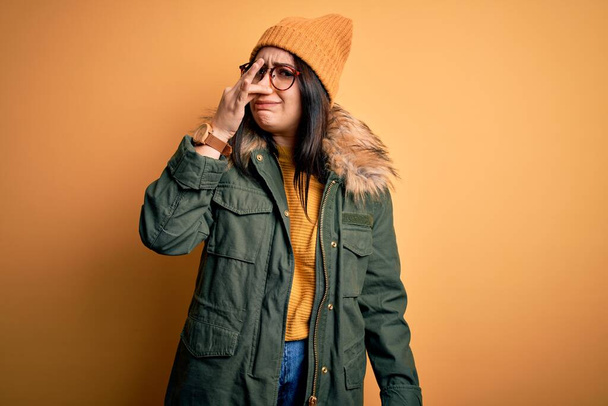 若いブルネットの女性は、黄色の隔離された背景の上に帽子をかぶった眼鏡と冬のコートを身に着けている何か臭いと嫌な、耐え難い匂いを嗅ぎ、鼻の上に指で息を保持する。匂いが悪い - 写真・画像