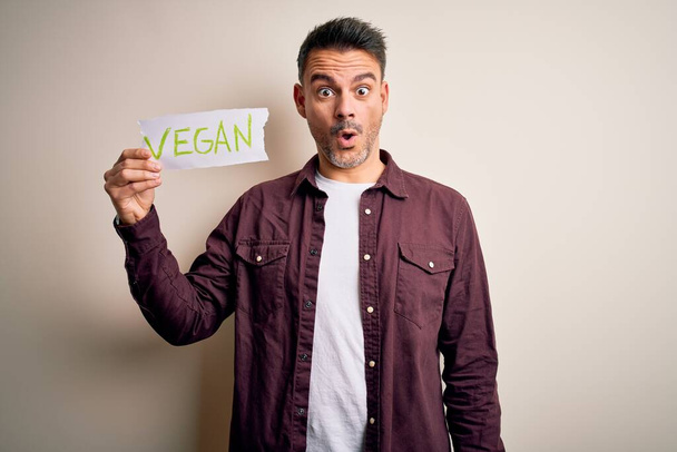 Jonge knappe veggie man met papier met veganistische boodschap over witte achtergrond bang in shock met een verrassing gezicht, bang en opgewonden met angst uitdrukking - Foto, afbeelding