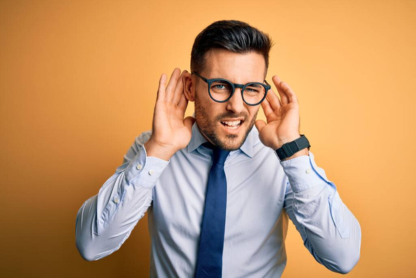 Молодой привлекательный бизнесмен в галстуке и очках стоит на жёлтом фоне, пытаясь услышать жесты обеих рук, любопытные для сплетен. Проблемы со слухом, глухой
 - Фото, изображение