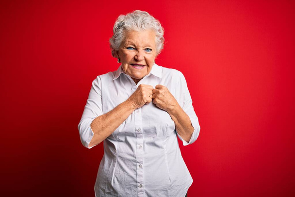 Senior mooie vrouw dragen elegante shirt staan over geïsoleerde rode achtergrond lachen nerveus en opgewonden met handen op kin kijken naar de zijkant - Foto, afbeelding