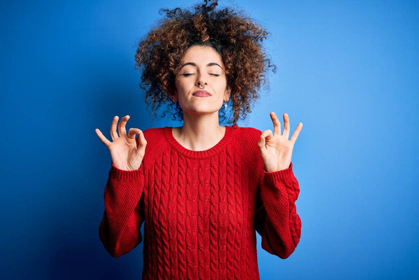 Young beautiful woman with curly hair and piercing wearing casual red sweater relax and smiling with eyes closed doing meditation gesture with fingers. Yoga concept. - Fotoğraf, Görsel